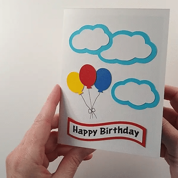Hands opening an intricate sliceform card featuring a colorful pop-up hot air balloon, birthday balloons and clouds.