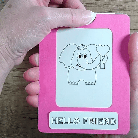 Hand pulling tab on a pink framed Magic slider card to change a black & white elephant to color with a 'Hello Friend' message